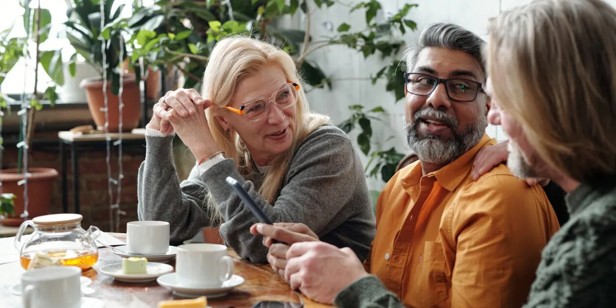 group of mature friends gathering in cozy cafe, enjoying coffee and pastries while discussing ideas hanging out in relaxed, friendly atmosphere with plants and sunlight streaming in