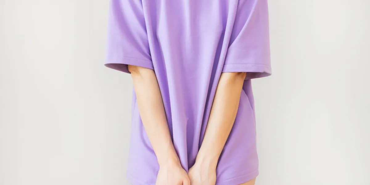 close-up of a young woman standing in a purple t-shirt and covering her crotch with her hands, isolated on a beige background suffers from urinary incontinence, cystitis, shy gynecological problems the concept of women's health