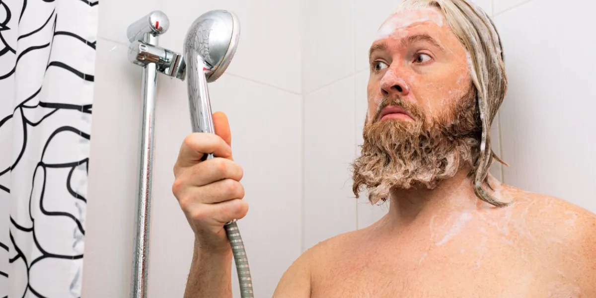 confused bearded man with soapy head standing in the bathroom and looking at the shower while the water supply has stopped no water coming from tap