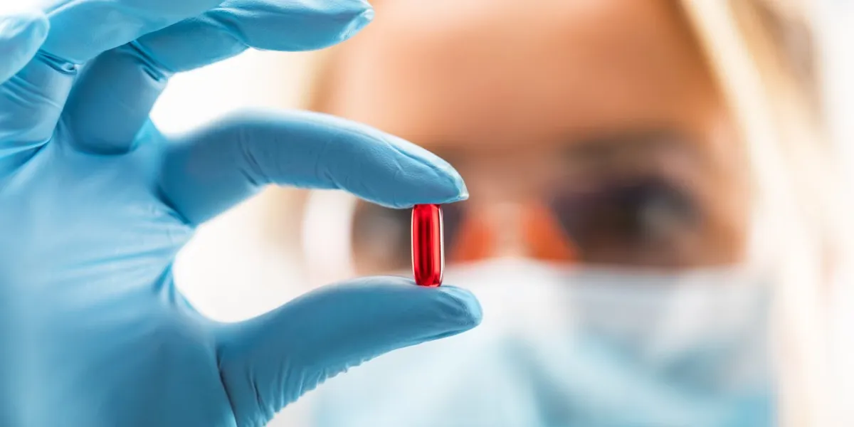 young attractive female scientist with protective eyeglasses and mask holding a red transparent pill with fingers in gloves in the pharmaceutical research laboratory