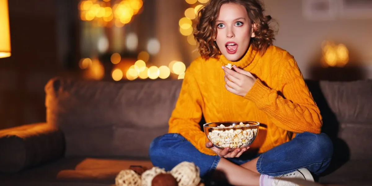 fascinated young female in sweater eating popcorn and watching interesting film on tv while sitting on couch in evening at home