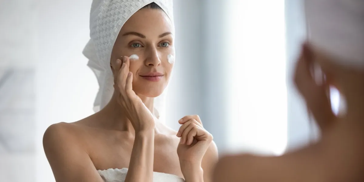 attractive young adult woman apply facial cream look in mirror, beautiful healthy lady wrapped in towels put moisturizing lifting nourishing day creme on soft hydrated moisturized skin in bathroom