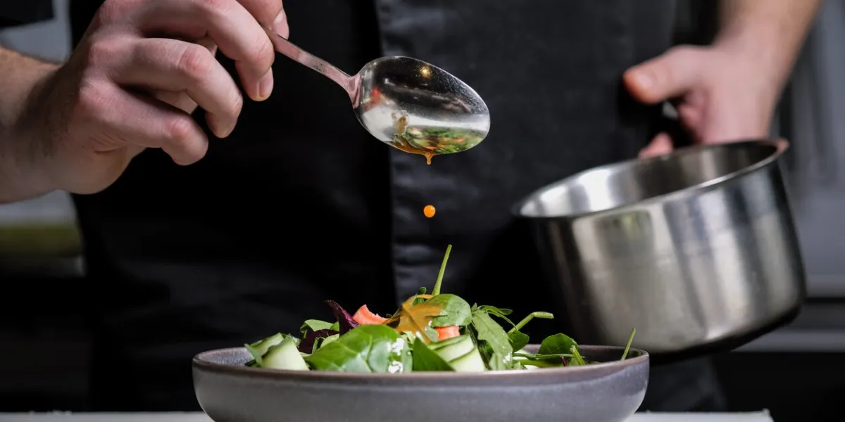 close-up of the hands of a male chef on a black background pour sauce from the spoon on the salad dish food decoration