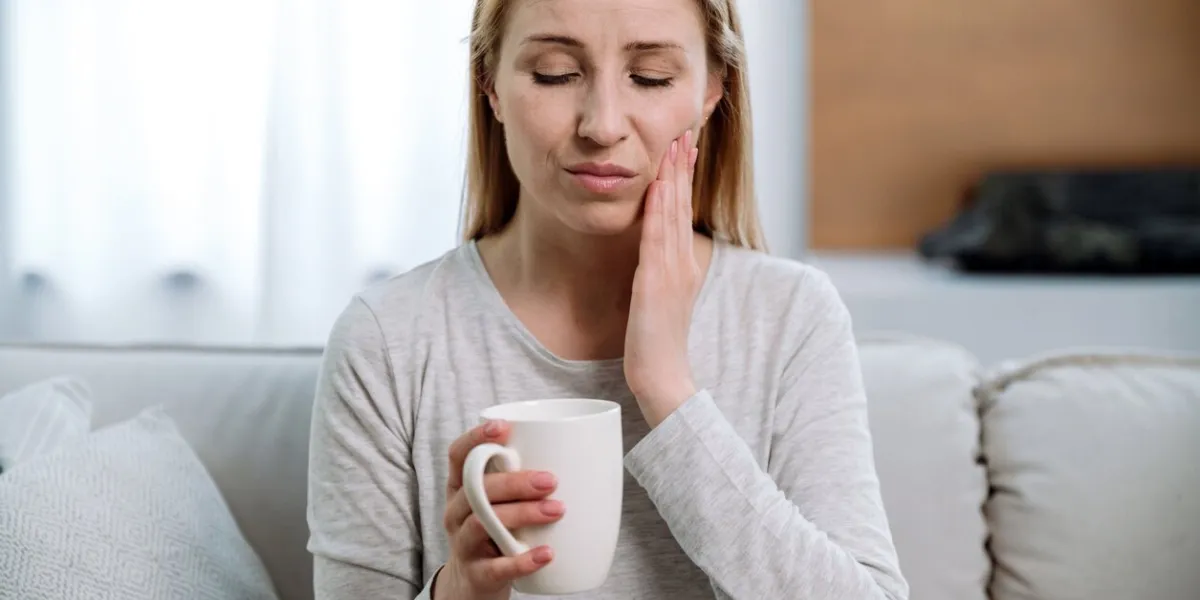 portrait of sad woman sitting on sofa, holding cup, feeling pain while drinking hot tea or cold beverage sensitive teeth dental and oral care for problem enamel