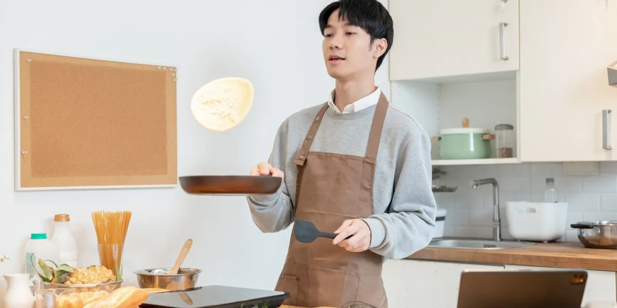a joyful and handsome young asian man in an apron, tossing pancakes on a frying pan, enjoys cooking pancakes in his kitchen