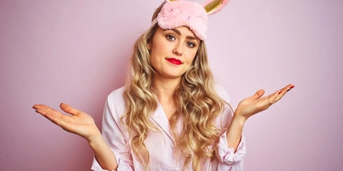 young beautiful woman wearing pajama and sleep mask over pink isolated background clueless and confused expression with arms and hands raised doubt concept