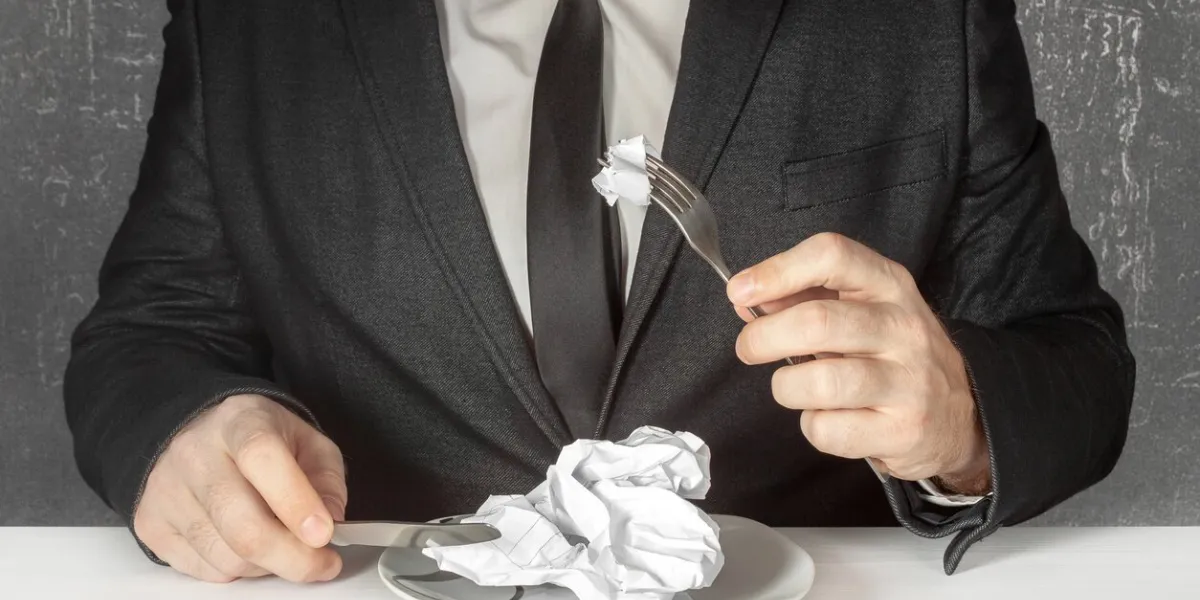 a man without a face in a black suit and tie sits at a table and prepares to eat a piece of a sheet of paper, a contract, an agreement, a table, financial documents using a knife and a fork destruction of compromising evidence
