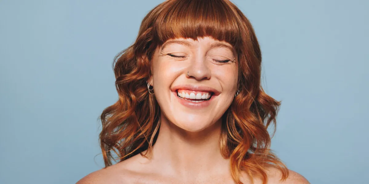 happy woman with ginger hair and flawless skin smiling with her eyes closed cheerful young woman embracing her natural beauty body confident young woman standing against a blue studio background