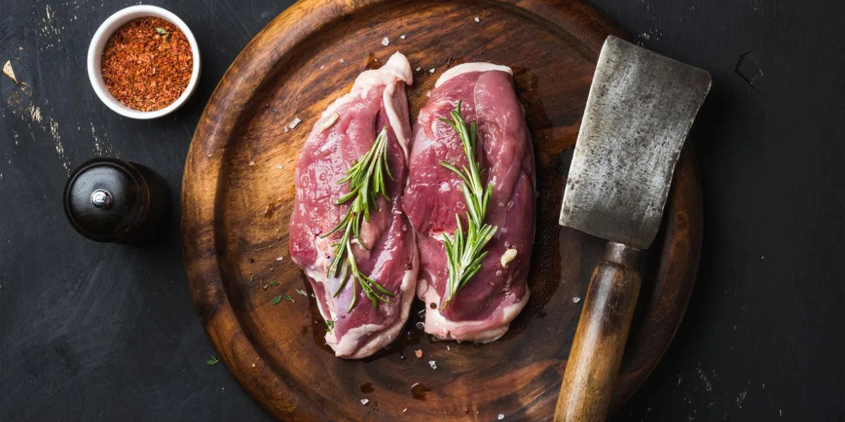 viande de volaille crue crue coupe poitrine de canard au romarin, épices et couperet boucher sur un plateau en bois foncé sur fond en bois noir, vue de dessus