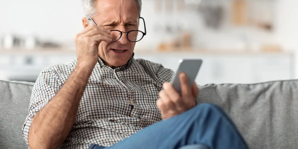 poor eyesight senior man squinting eyes reading message on phone wearing eyeglasses having problems with vision sitting on couch at home ophtalmic issue, bad sight in older age concept