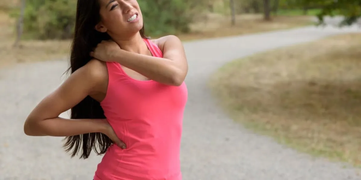 une jeune femme en train de faire du jogging subit une blessure musculaire debout en se tenant le cou et le bas du dos tout en grimaçant de douleur sur une route rurale, près du haut du corps
