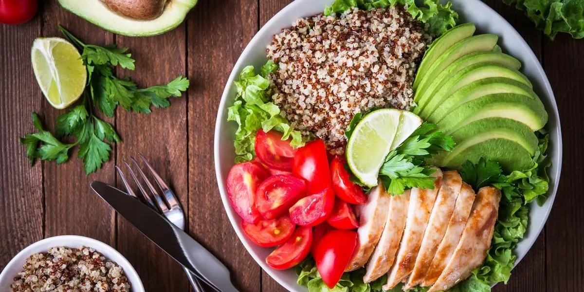 healthy salad bowl with quinoa, tomatoes, chicken, avocado, lime and mixed greens (lettuce, parsley) on wooden background top view food and health