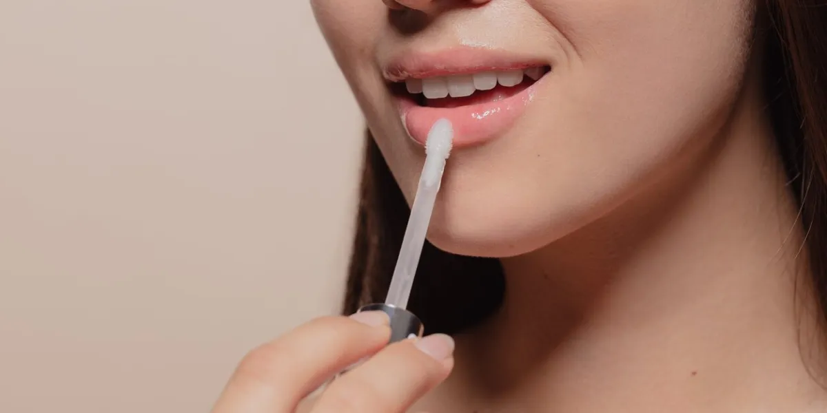 close up of young korean woman applying transparent lip gloss cropped shot of girl putting on makeup on her lips with applicator against beige background