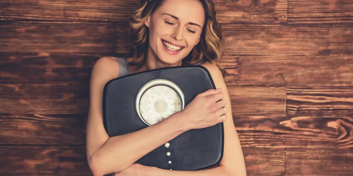 beautiful young woman in sportswear is hugging weigh scales and smiling, on wooden background