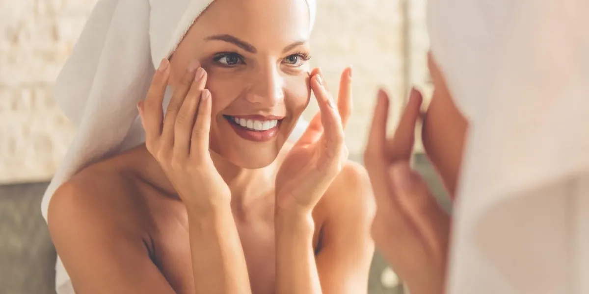 belle jeune femme en serviette de bain touche son visage et souriant tout en regardant dans le miroir dans la salle de bain