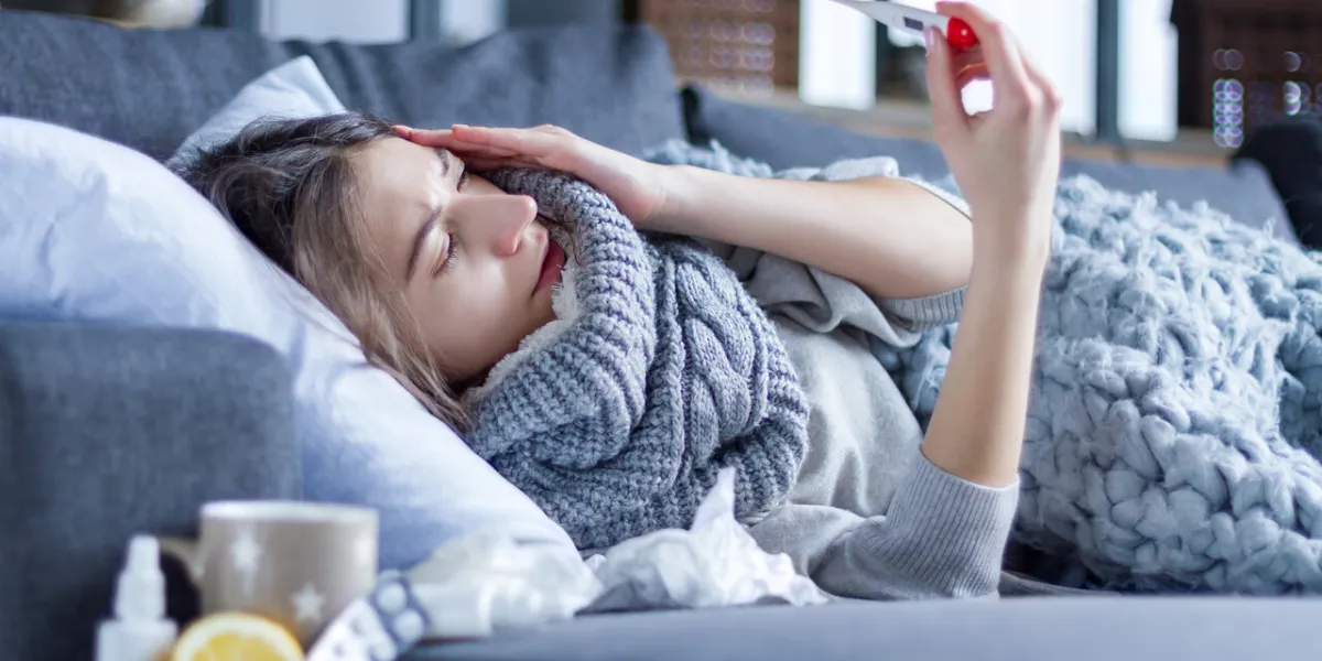 sick exhausted girl in scarf is lying in bed wrapped in blanket young woman with fever and headache is measuring temperature with thermometer, treated at home winter cold and flu concept