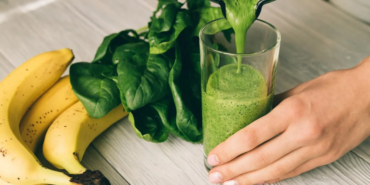 female hand pours a smoothie of banana and spinach in glass on a wooden table