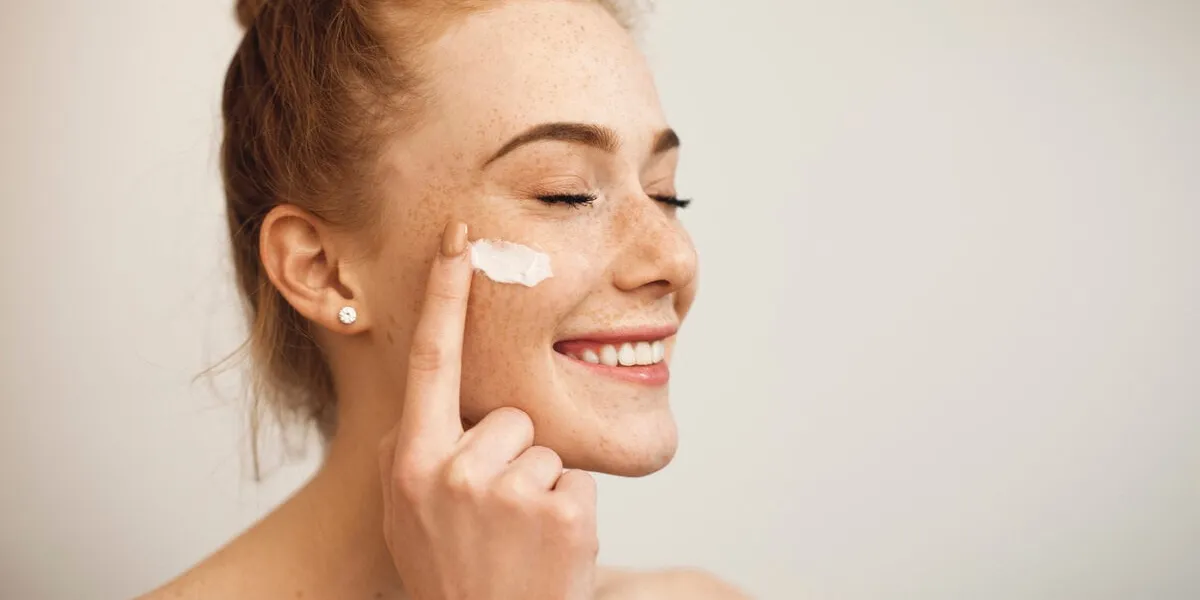 close up of a young female with red hair and freckles applying white cream on her face laughing with closed eyes isolated on white background