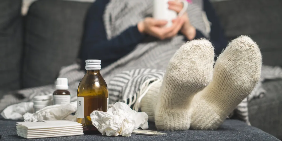 cold medicine and sick woman drinking hot beverage to get well from flu, fever and virus dirty paper towels and tissues on table ill person wearing warm woolen stocking socks in winter