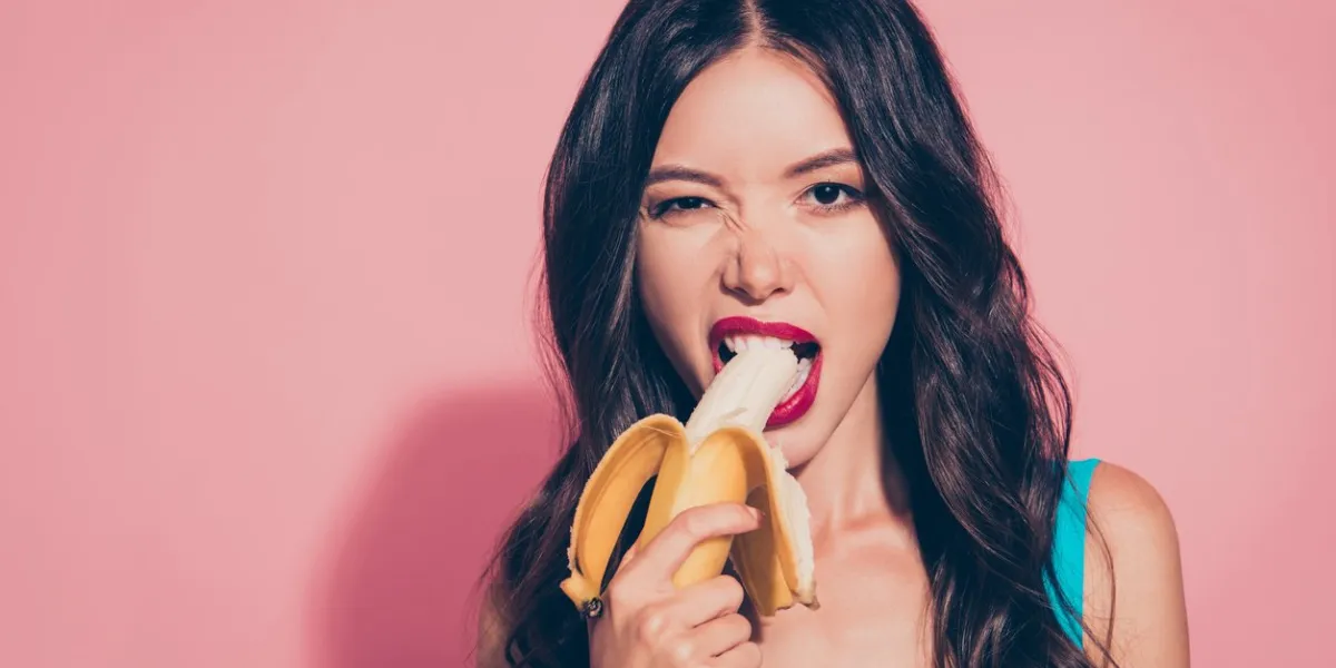close-up portrait of nice cheeky cunning adorable glamorous charming attractive wavy-haired lady holding biting banana red lips isolated over pink pastel background
