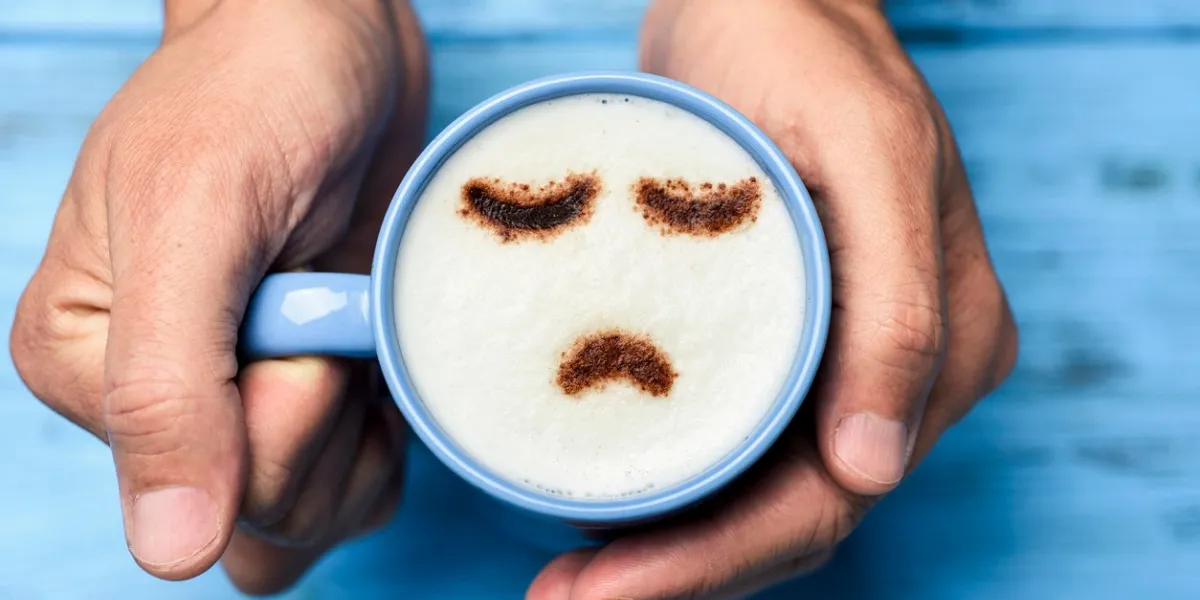 high-angle shot of a young caucasian man with a blue cup of cappuccino with a sad face drawn with cocoa powder on the milk foam, on a blue rustic table