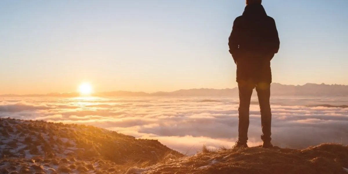 view from the back a lonely standing man high in the mountains looks at the setting sun and the sunset horizon with a valley filled with clouds the concept of tourism travel and male loneliness