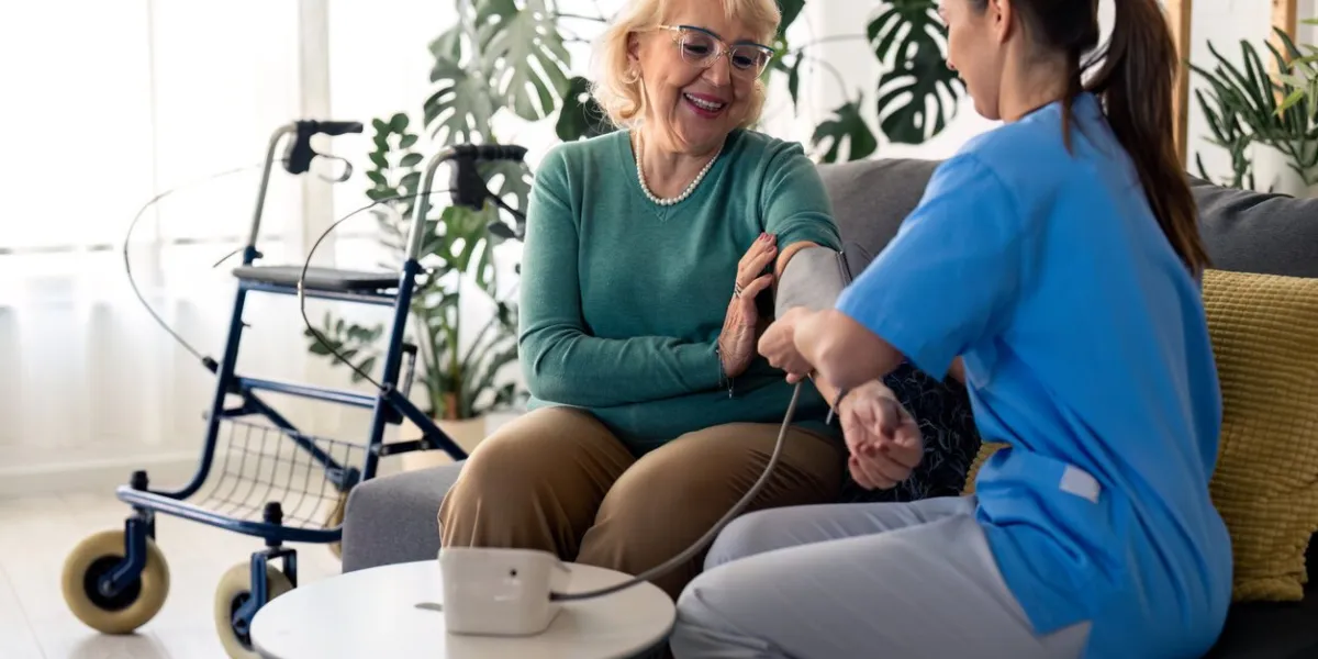 beautiful senior woman measuring blood pressure with help of home nurse, sitting on sofa in living room, feeling satisfied and happy with home care female nurse assisting senior woman to measure blood pressure