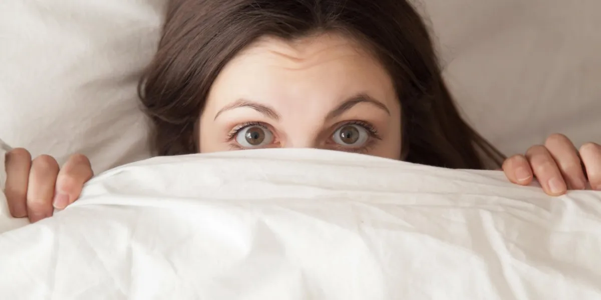 funny surprised girl covering half of face with white blanket, young scared woman hiding and peeking from duvet, afraid of night monsters, feels embarrassed, wide awake, head shot close up, top view