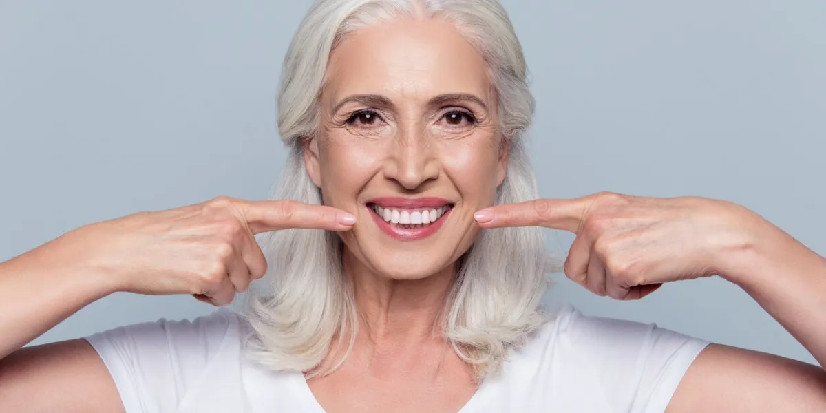 concept of having strong healthy straight white teeth at old age close up portrait of happy with beaming smile female pensioner pointing on her perfect clear white teeth, isolated on gray background