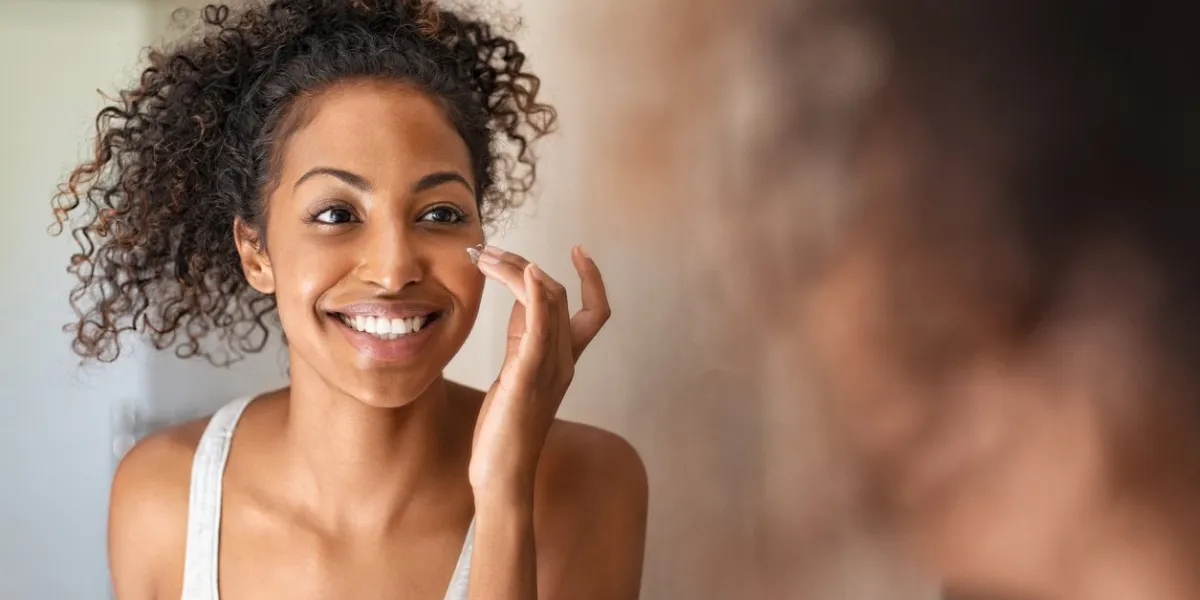 young black woman applying moisturising cream to her skin while standing in front of the mirror in the bathroom african american girl applying face cream while smiling beauty hydrating moisturizer and skincare routine concept
