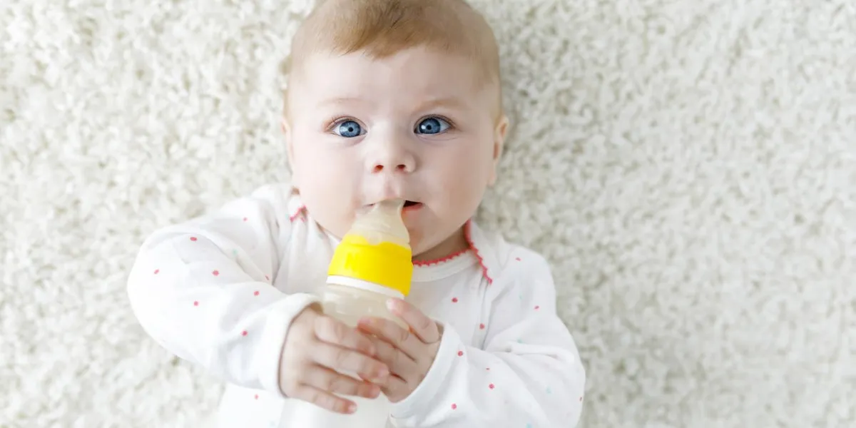 cute adorable newborn baby girl holding nursing bottle and drinking formula milk first food for babies new born child, little girl laying on white background family, new life, childhood, bottle-feeding concept