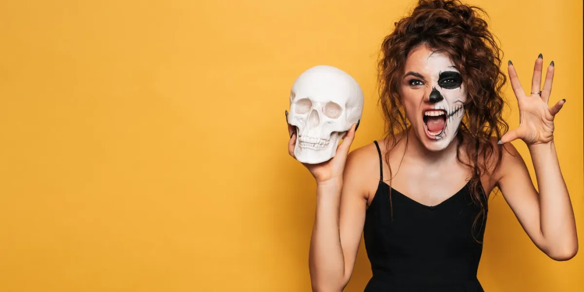 a young woman in a halloween costume with makeup for the holiday on a yellow isolated background with a skull in her hands the emotion on the face is fear-inducing a place for your text or advertisement