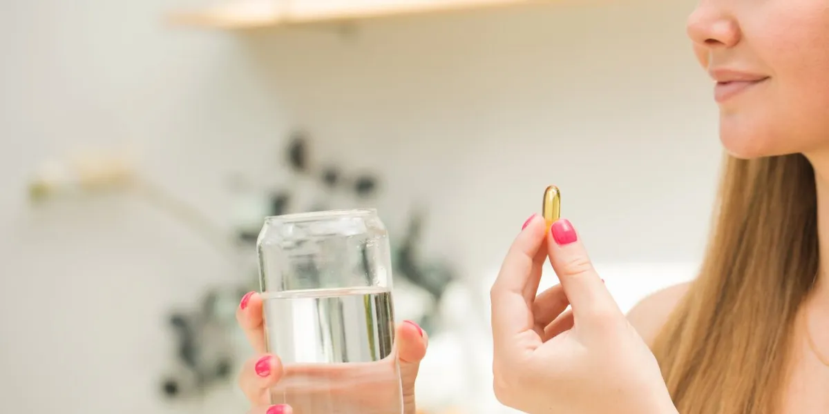happy blonde woman holding a glass of water and a capsule of fish oil or vitamin d in the kitchen vitamins and nutritional supplements healthy lifestyle preventive medicine