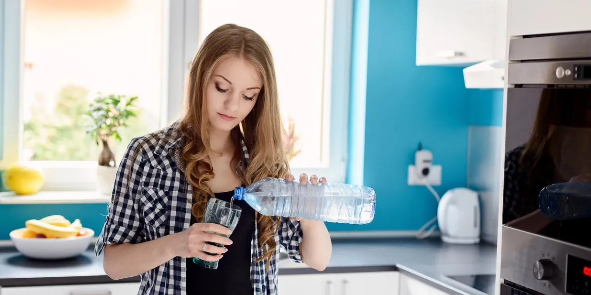 belle femme blonde verser de l'eau d'une bouteille dans un verre debout dans la cuisine, vêtue d'une chemise à carreaux