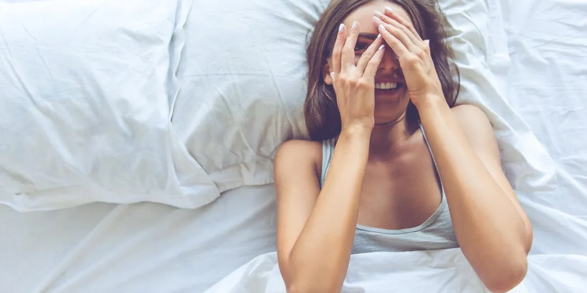 top view of beautiful sexy young woman covering her eyes, looking at camera and smiling while lying in the bed
