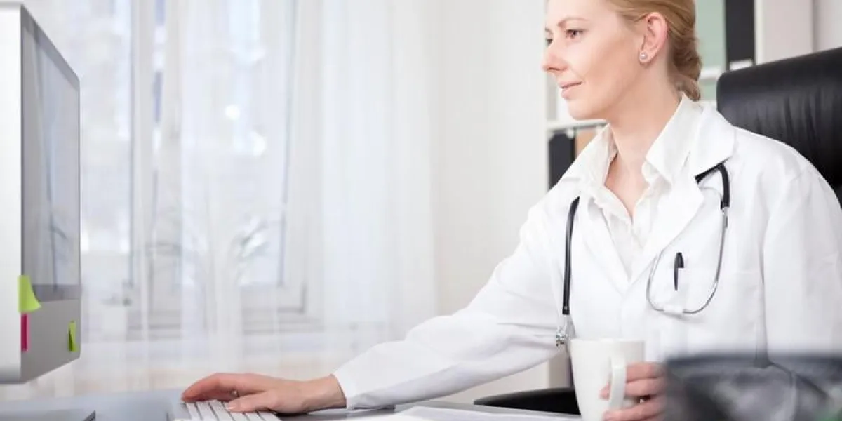 souriant adulte femme médecin à son bureau de surfer sur internet en utilisant son ordinateur de bureau tout en prenant une tasse de boisson
