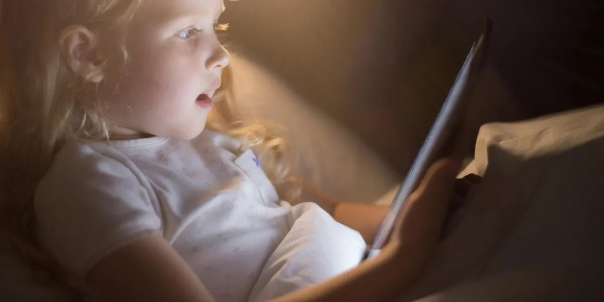 side view portrait of adorable little girl looking amazed with mouth open using digital tablet lying in bed at night, face lit by screen light like magic, copy space