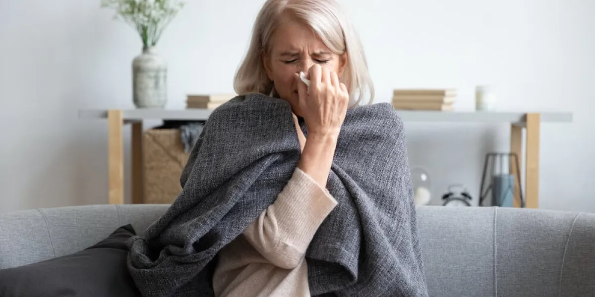 middle-aged 50s sick frozen woman seated on sofa in living room covered with warm plaid sneezing holding paper napkin blow out runny nose feels unhealthy, seasonal cold, weakened immune system concept