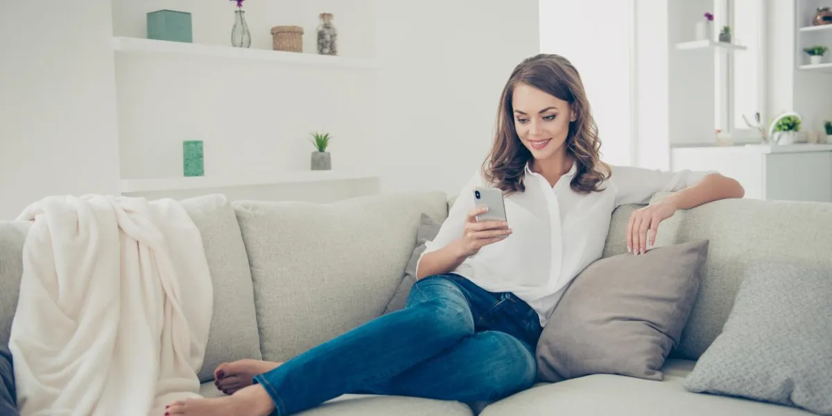 portrait of pretty, charming, cheerful, trendy house wife in jeans, shirt with curly hair sitting on sofa having cell smart phone in hands chatting with lover searching contact using 3g wi-fi internet