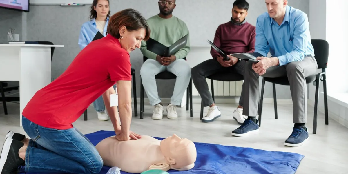medical instructor showing cpr on training mannequin on floor to people who sitting on chairs and watching during lesson