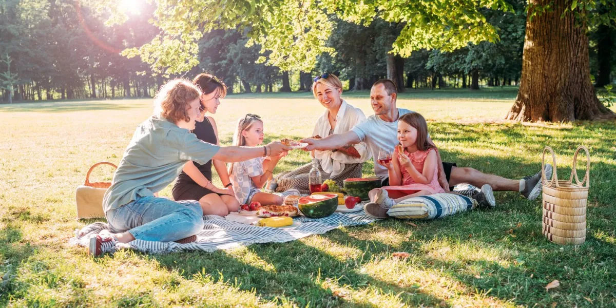 big family sitting on the picnic blanket in city park during weekend sunny day they smiling, laughing and eating watermelon, boiled corn,pie with cold tea family values, outdoors activities concept