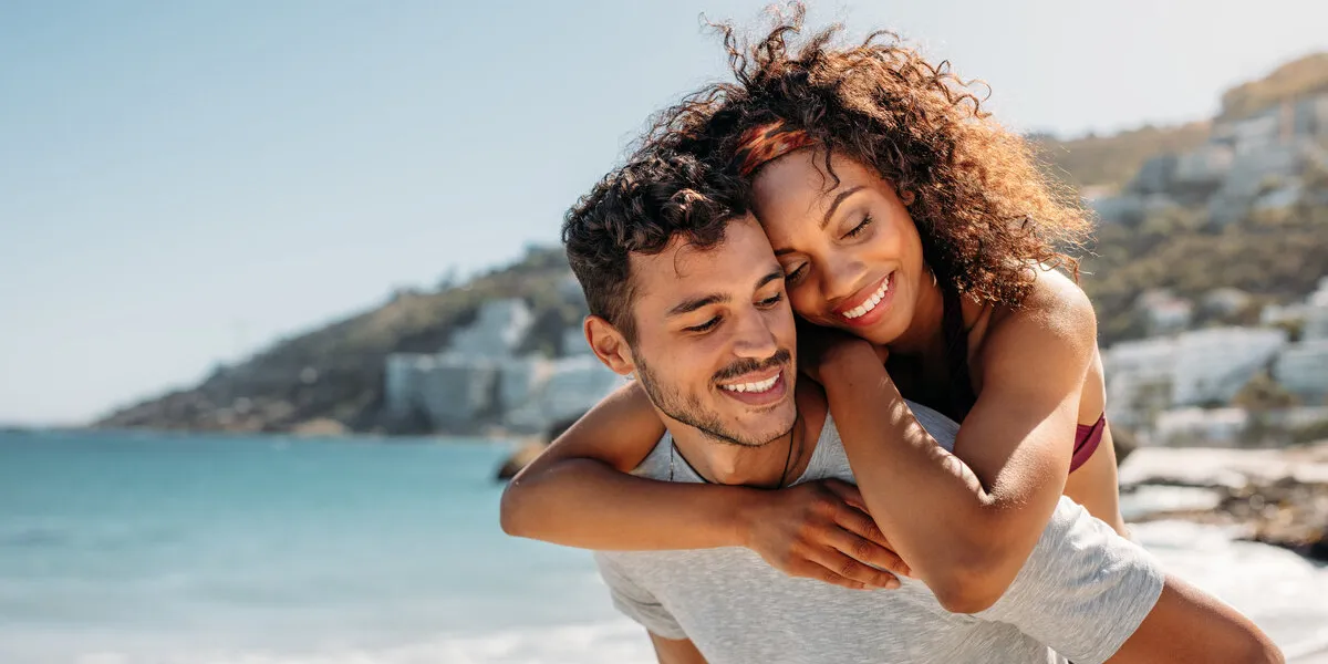 happy man carrying a woman on his back walking on beach with sea in the background tourist couple on vacation spending time together on the beach