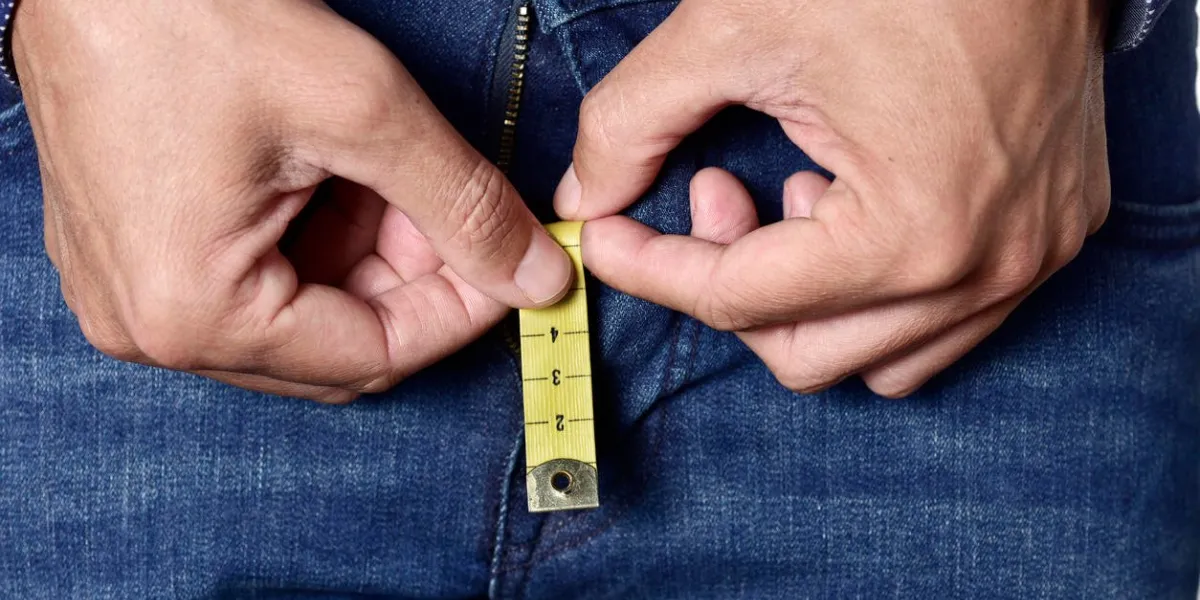 closeup of a young man holding a short piece of measuring tape that is popping up from the fly of his jeans