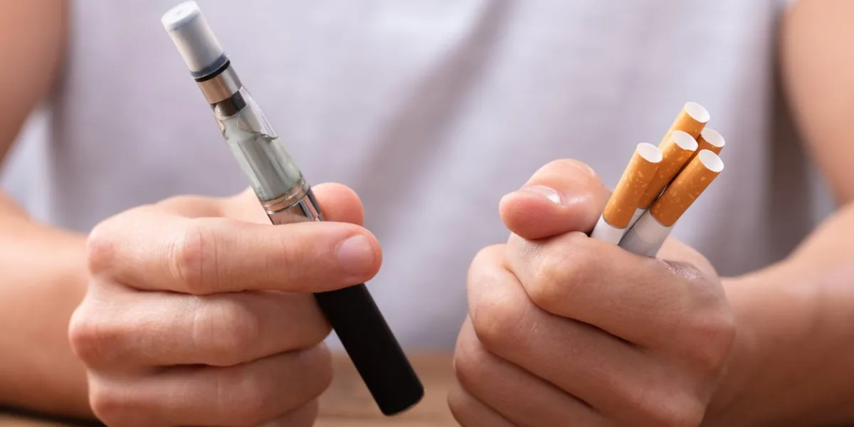 man holding vape and tobacco cigarette over desk