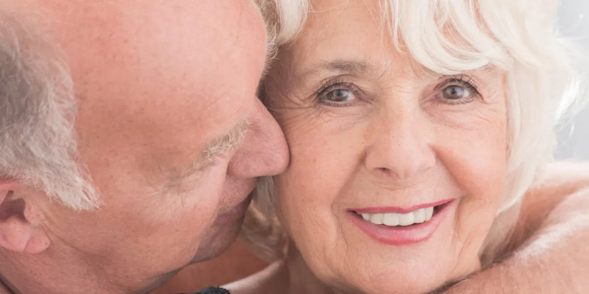 cropped picture of a senior woman smiling at the camera and her husband kissing her on the cheek