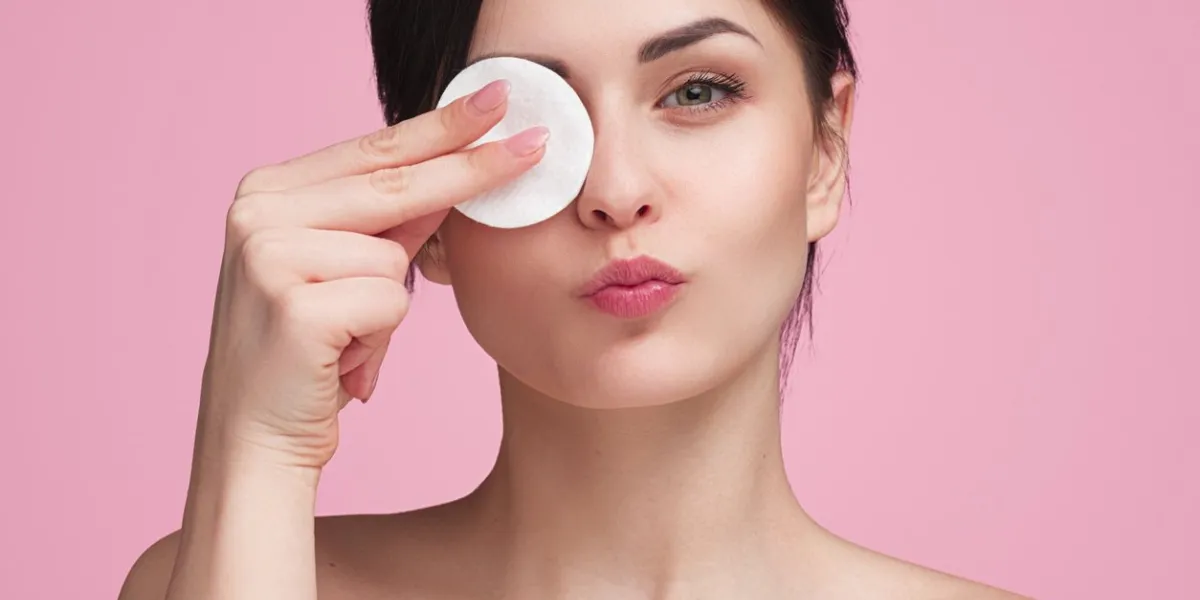 pretty young female wiping eye with cotton pad in studio with pink background and looking at camera