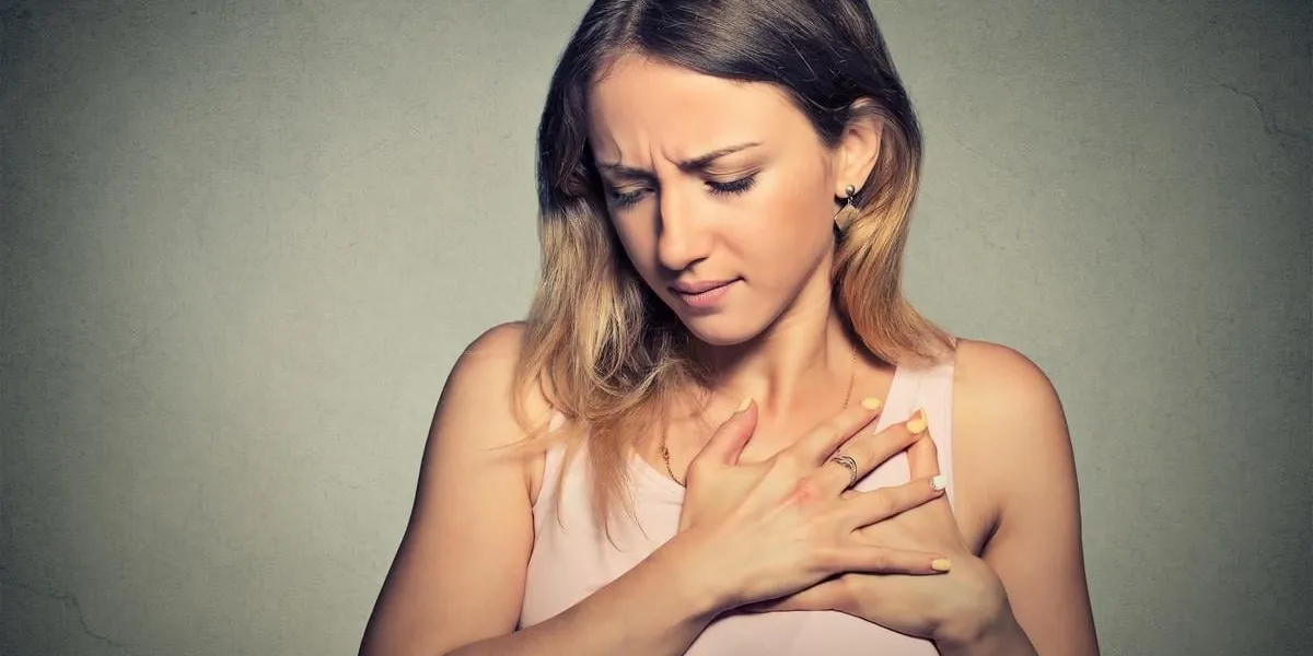 sick woman with heart attack, pain, health problem holding touching her chest with hands isolated on gray wall background human face expression