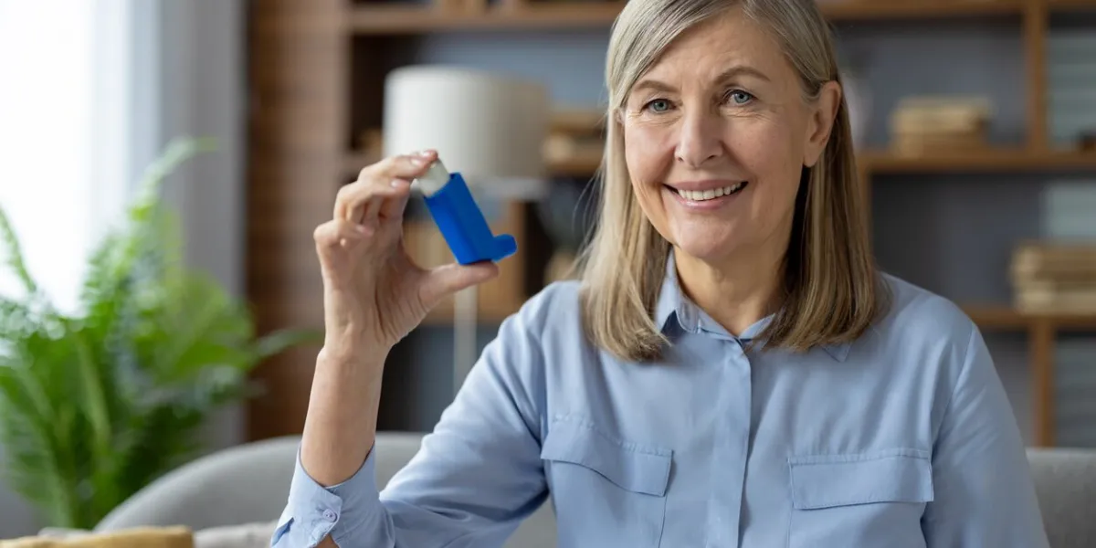 smiling woman with blue inhaler in hand looking at camera while sitting in domestic atmosphere wrinkled female getting used to daily routine for health and wellness with chronically disease