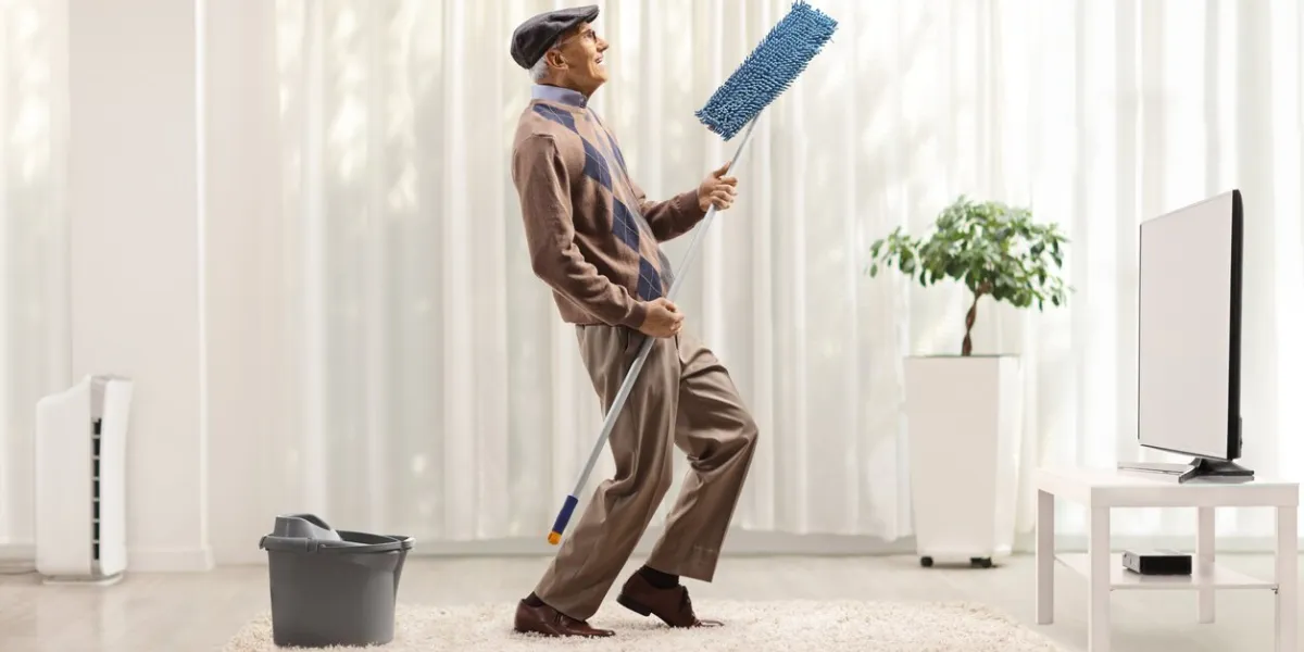 elderly man singing and dancing with a cleaning mop at home in front of tv in a living room