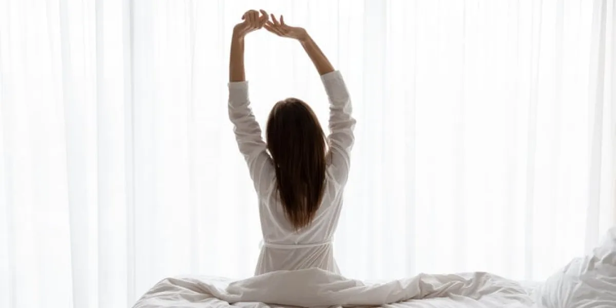 rear view beautiful woman wearing white bathrobe doing exercise, sitting on comfortable bed in modern apartment or hotel, young female stretching arms in early morning after awakening